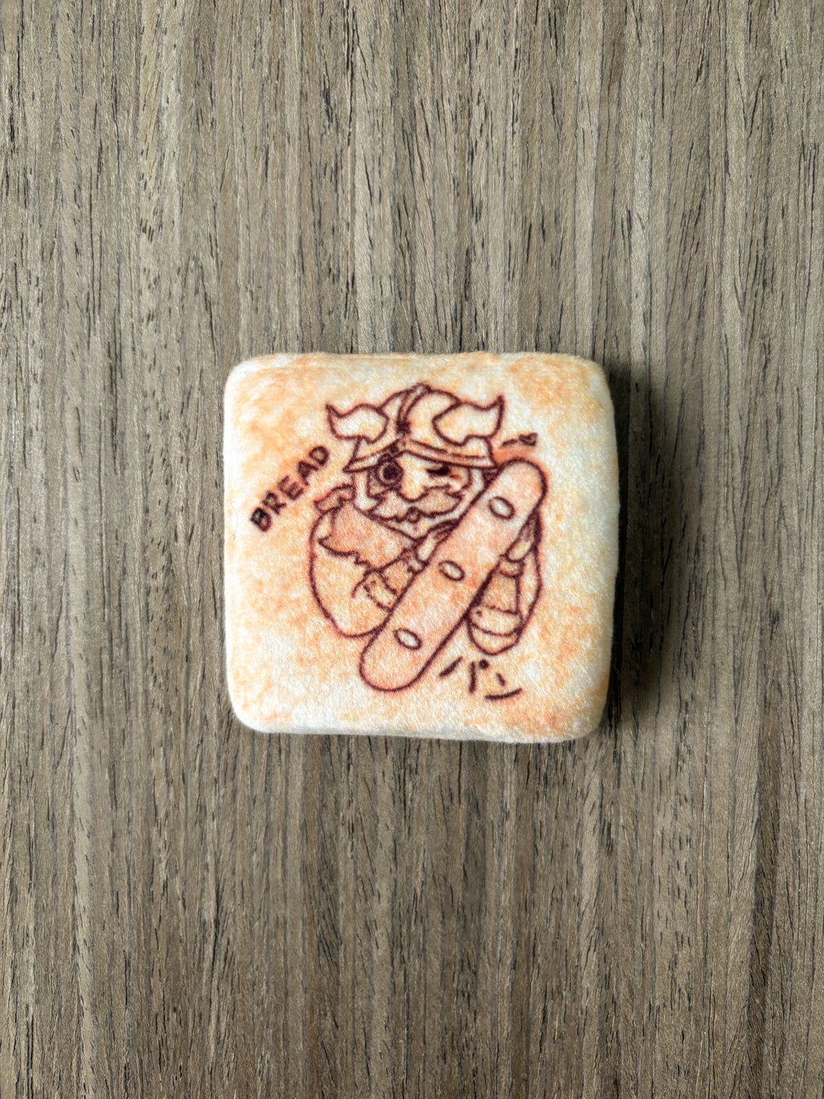 A plushie tin badge resembling a piece of bread with a Senshi from Dungeon Meshi "toasted" on it.