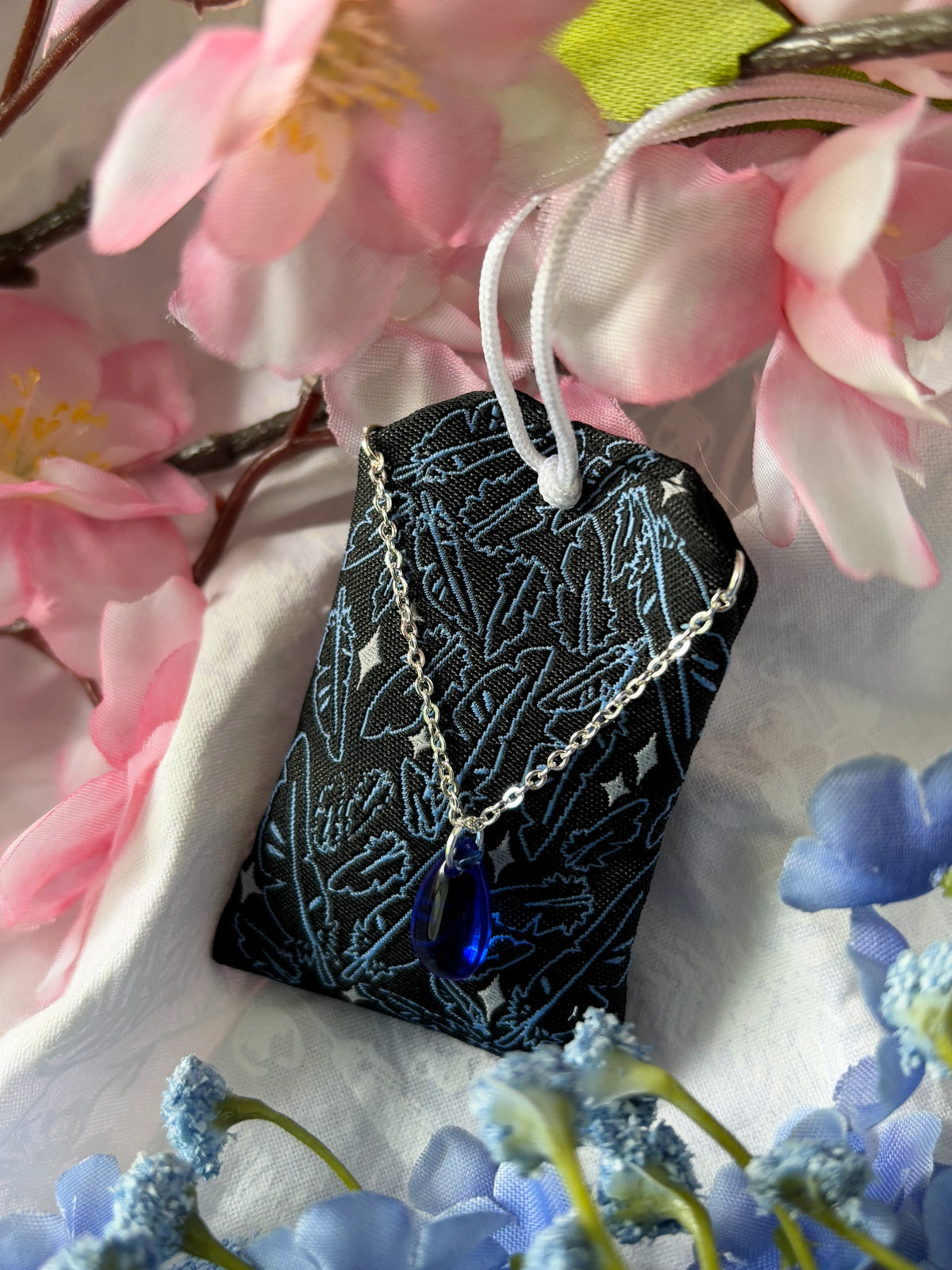 Japanese good luck charm (omamori) based on the Studio Ghibli movie Howl's Moving Castle. This is a black, blue, and grey fabric charm that displays Howl's black feathers (Howl in his monster form) alongside Sophie's starlight hair braid. The back of the charm displays Howl's feathers with his blue gem necklace.
