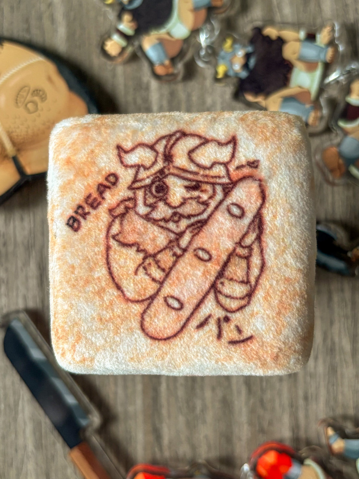 A plushie tin badge resembling a piece of bread with a Senshi from Dungeon Meshi "toasted" on it.
