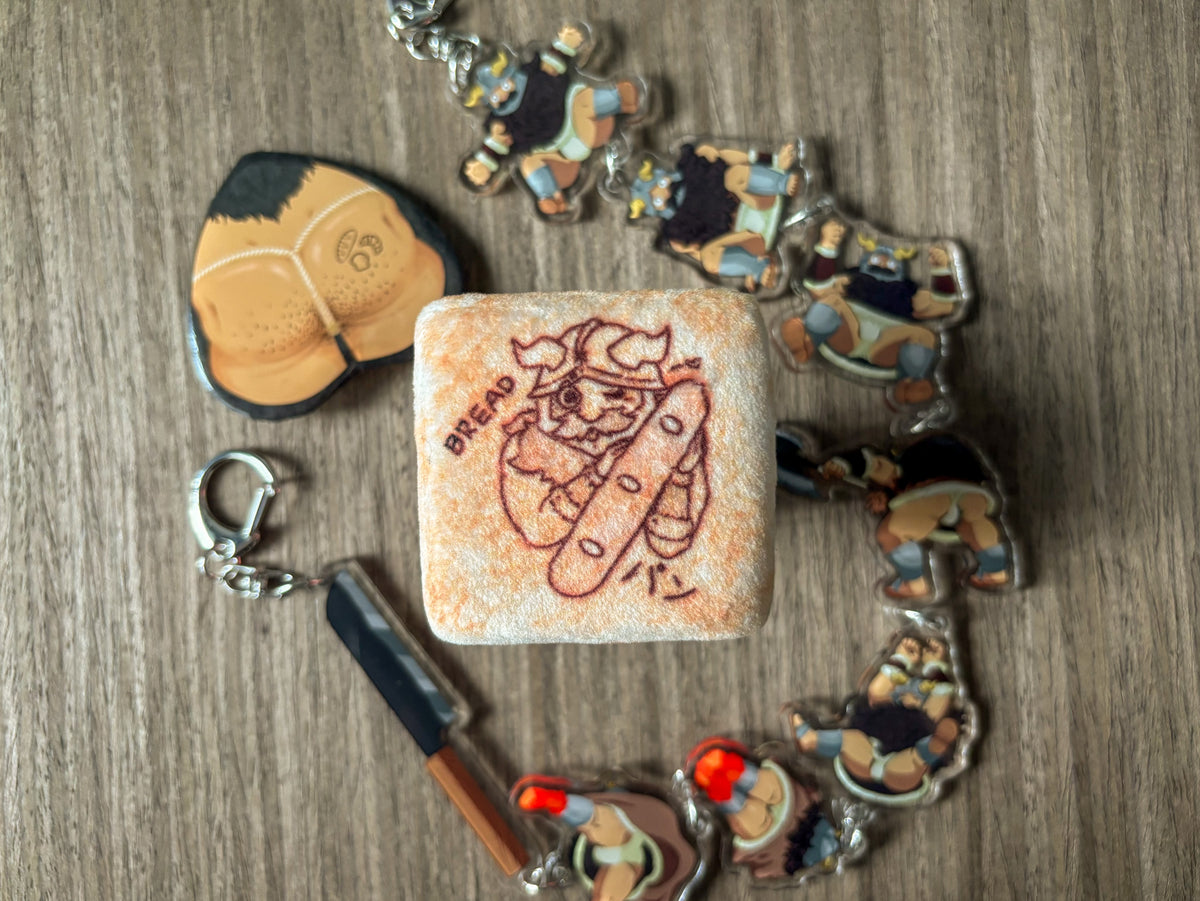 A plushie tin badge resembling a piece of bread with a Senshi from Dungeon Meshi "toasted" on it.