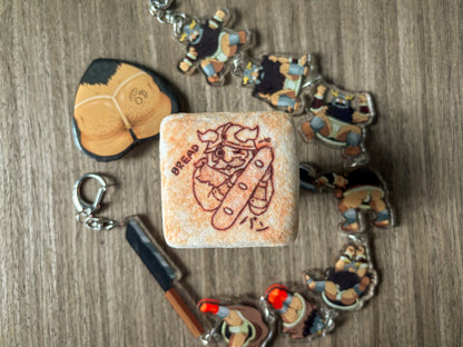 A plushie tin badge resembling a piece of bread with a Senshi from Dungeon Meshi "toasted" on it.