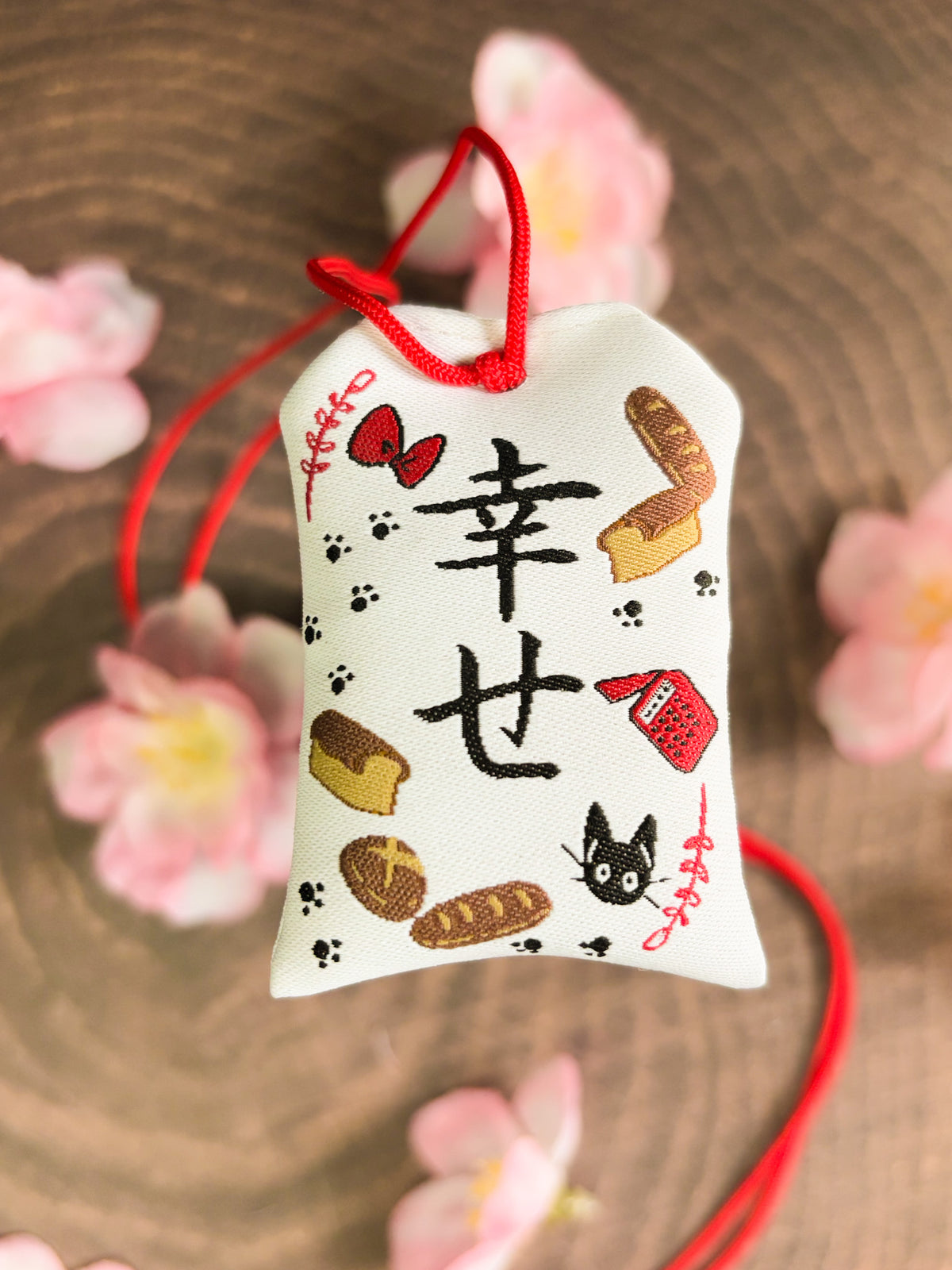 Japanese good luck charm (omamori) based on the Studio Ghibli movie Kiki's Delivery Service. This is a mostly white fabric charm that displays items from Kiki's Delivery Service such as Jiji, Kiki's bow, bakery goods, and her broom.