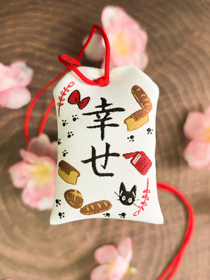 Japanese good luck charm (omamori) based on the Studio Ghibli movie Kiki's Delivery Service. This is a mostly white fabric charm that displays items from Kiki's Delivery Service such as Jiji, Kiki's bow, bakery goods, and her broom.