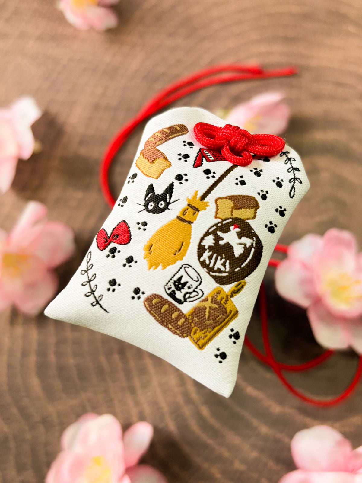 Japanese good luck charm (omamori) based on the Studio Ghibli movie Kiki's Delivery Service. This is a mostly white fabric charm that displays items from Kiki's Delivery Service such as Jiji, Kiki's bow, bakery goods, and her broom.