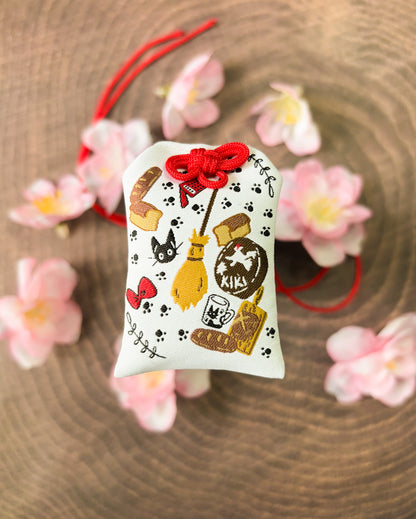Japanese good luck charm (omamori) based on the Studio Ghibli movie Kiki's Delivery Service. This is a mostly white fabric charm that displays items from Kiki's Delivery Service such as Jiji, Kiki's bow, bakery goods, and her broom.