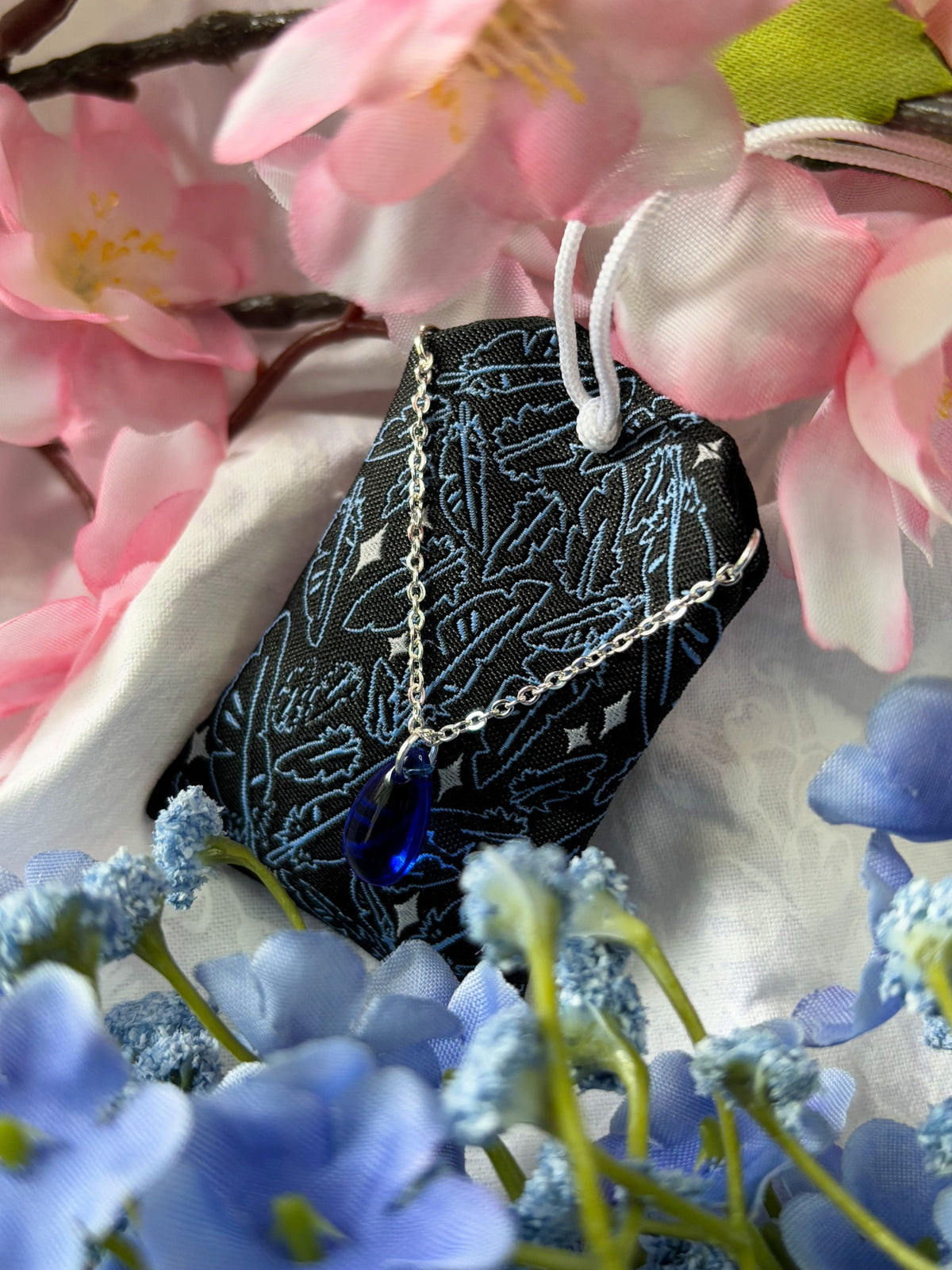 Japanese good luck charm (omamori) based on the Studio Ghibli movie Howl's Moving Castle. This is a black, blue, and grey fabric charm that displays Howl's black feathers (Howl in his monster form) alongside Sophie's starlight hair braid. The back of the charm displays Howl's feathers with his blue gem necklace.