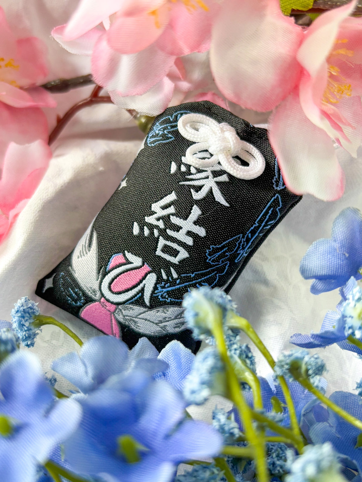 Japanese good luck charm (omamori) based on the Studio Ghibli movie Howl's Moving Castle. This is a black, blue, and grey fabric charm that displays Howl's black feathers (Howl in his monster form) alongside Sophie's starlight hair braid. The back of the charm displays Howl's feathers with his blue gem necklace.