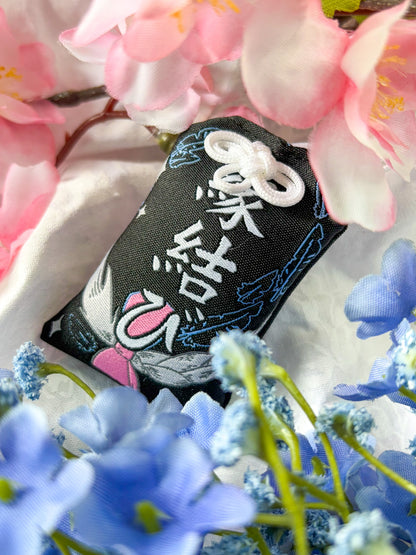 Japanese good luck charm (omamori) based on the Studio Ghibli movie Howl's Moving Castle. This is a black, blue, and grey fabric charm that displays Howl's black feathers (Howl in his monster form) alongside Sophie's starlight hair braid. The back of the charm displays Howl's feathers with his blue gem necklace.