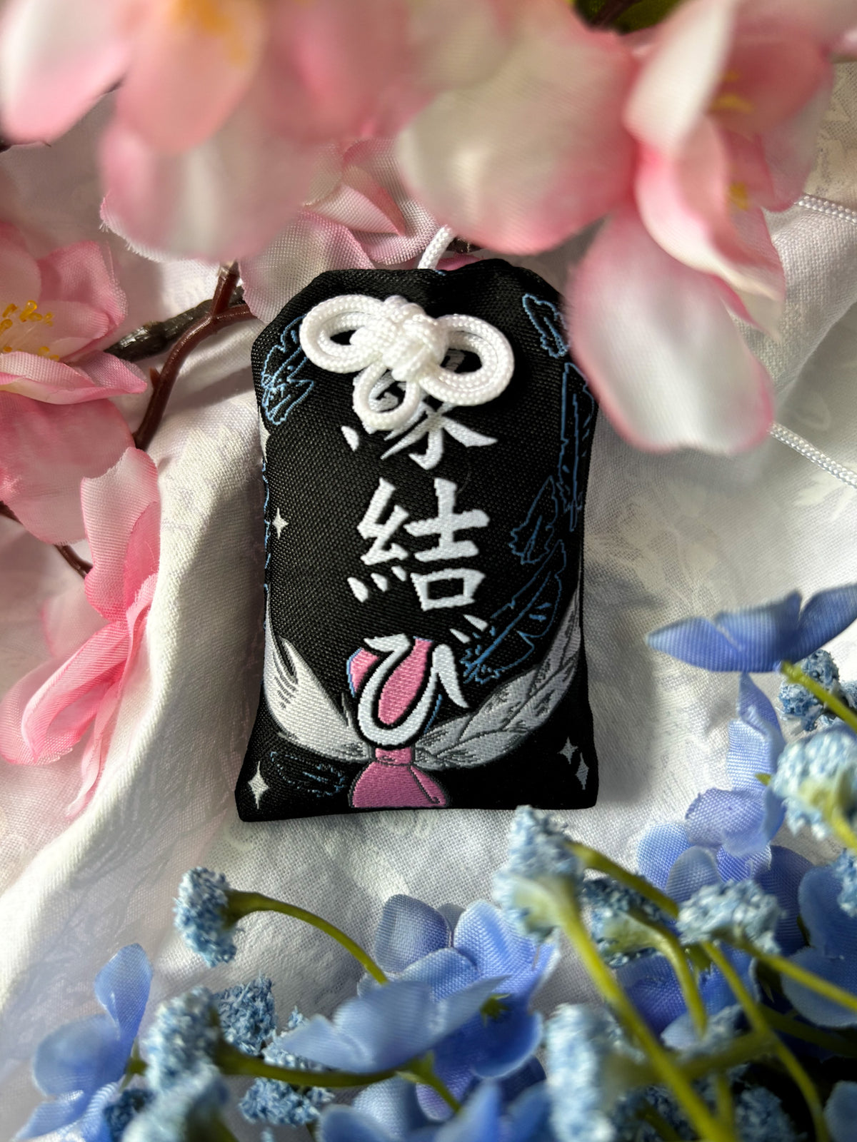 Japanese good luck charm (omamori) based on the Studio Ghibli movie Howl's Moving Castle. This is a black, blue, and grey fabric charm that displays Howl's black feathers (Howl in his monster form) alongside Sophie's starlight hair braid. The back of the charm displays Howl's feathers with his blue gem necklace.