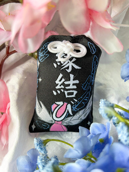 Japanese good luck charm (omamori) based on the Studio Ghibli movie Howl's Moving Castle. This is a black, blue, and grey fabric charm that displays Howl's black feathers (Howl in his monster form) alongside Sophie's starlight hair braid. The back of the charm displays Howl's feathers with his blue gem necklace.