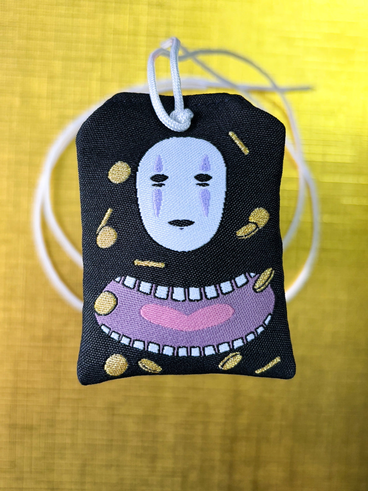 Japanese good luck charm (omamori) based on the Studio Ghibli movie Spirited Away. Based on the character No Face or Kaonashi. Charm displays No Face's mask with his mouth underneath with gold coins scattered around him. The front of the charm displays scattered and stacked coins.