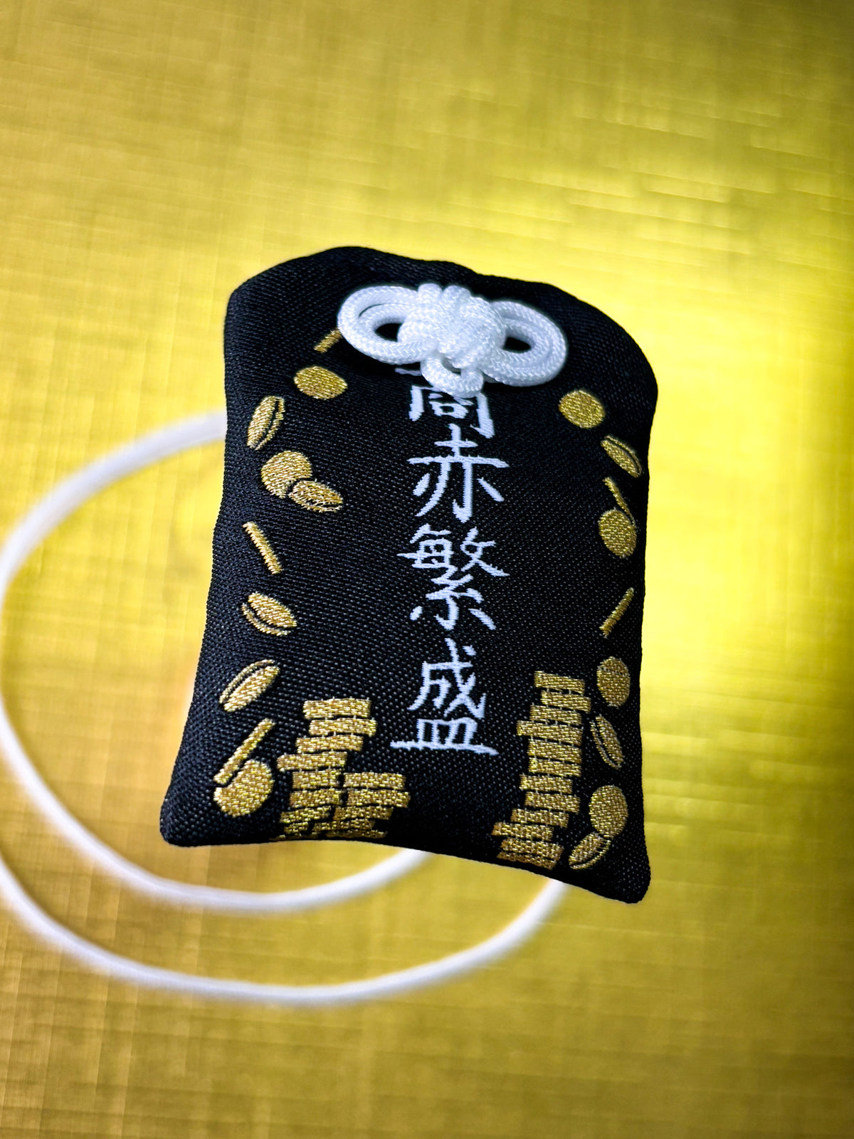 Japanese good luck charm (omamori) based on the Studio Ghibli movie Spirited Away. Based on the character No Face or Kaonashi. Charm displays No Face's mask with his mouth underneath with gold coins scattered around him. The front of the charm displays scattered and stacked coins.