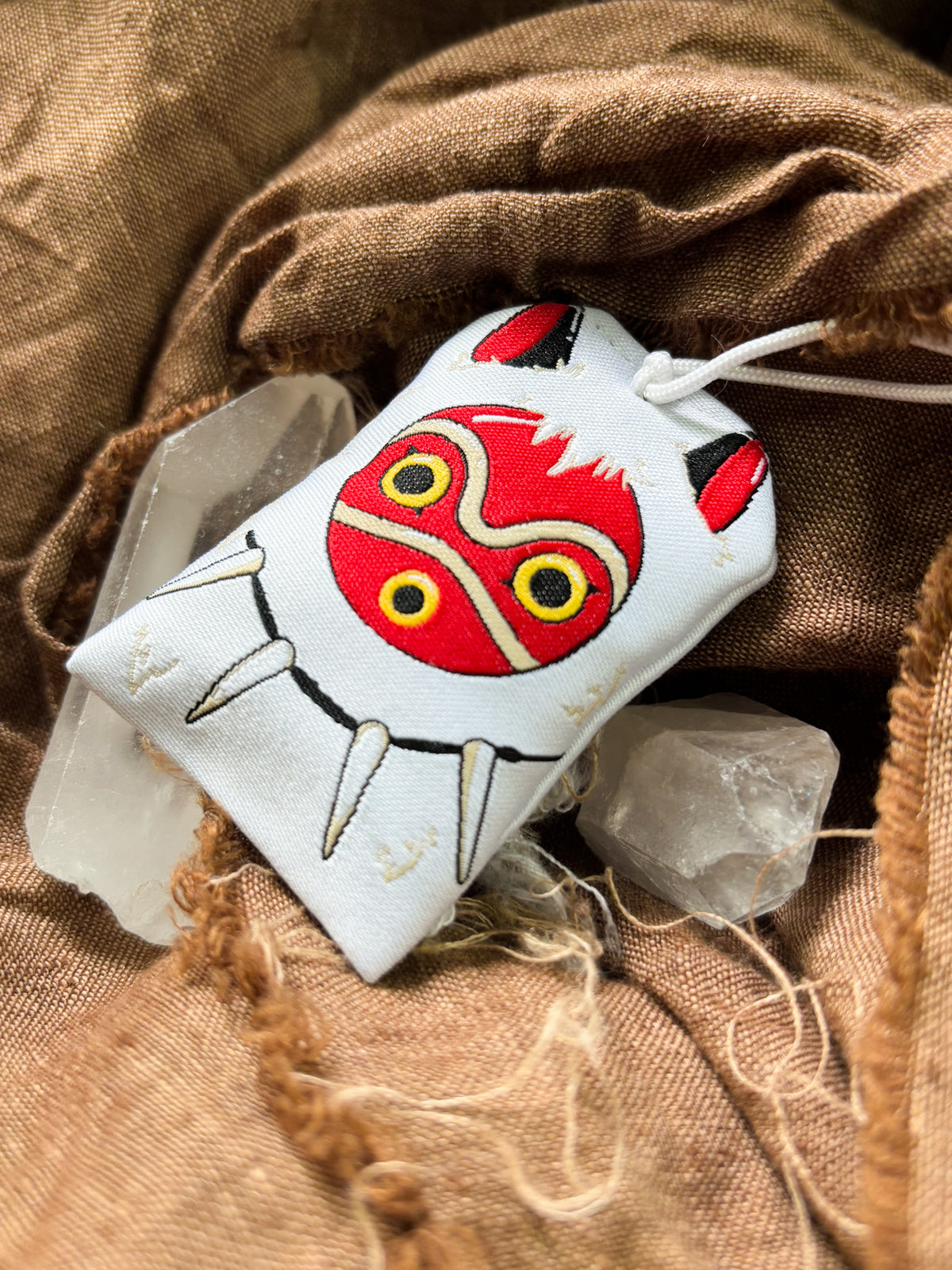 Japanese good luck charm (omamori) based on the Studio Ghibli movie Princess Mononoke. This is a mostly white fabric charm that displays San's mask and necklace. Charm replicates San's white fur cape. 